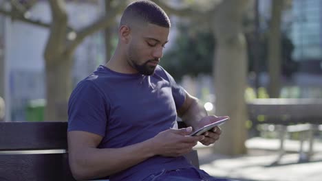 Afro-American-muscular-man-on-bench-in-park-typing-on-tablet